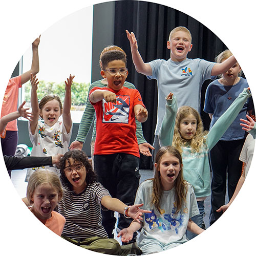 Photo from one of our kids performance classes. A group of kids are all singing in unison with great smiles and positive energy, holding their arms wide or pointing to the audience.