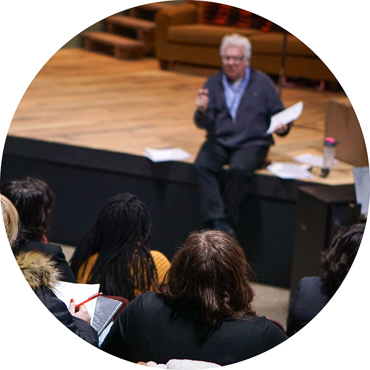 Photo from one of our audition workshops showing Dan Murphy sitting on the edge of the stage speaking with an audience of workshoppers listening.