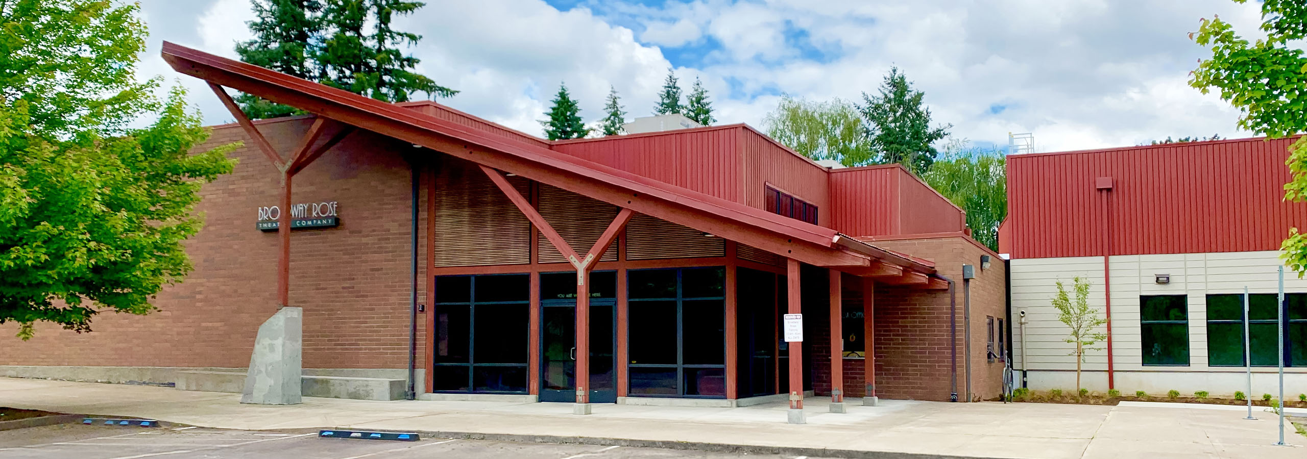 Photo of the front of the New Stage building. The roof over the atrium is sloped down and to the right. The front wall is all glass with a set of double doors leading into the building. The roof and support beams out front are red, but the building is mostly brown brick and beautiful wooden slats.