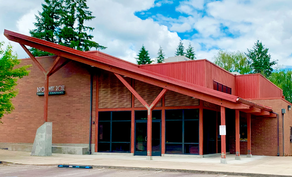 Photo of the front of the New Stage building. The roof over the atrium is sloped down and to the right. The front wall is all glass with a set of double doors leading into the building. The roof and support beams out front are red, but the building is mostly brown brick and beautiful wooden slats.