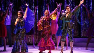Photo from our 2018 production of Mamma Mia, with three performers in glittering brightly colored disco outfits, singing together.
