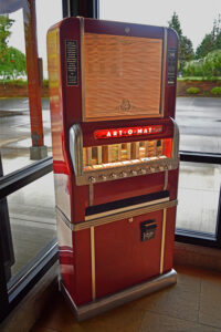 Photo of the Art-o-mat machine in the lobby. It stands about 5 and a half feet tall and is in a midcentury, 1950s industrial chic design. It is a bold cherry red with chrome accents and knobs. A small window in the front that used to show the cigarette brands inside now displays the artists' names and samples of their work.