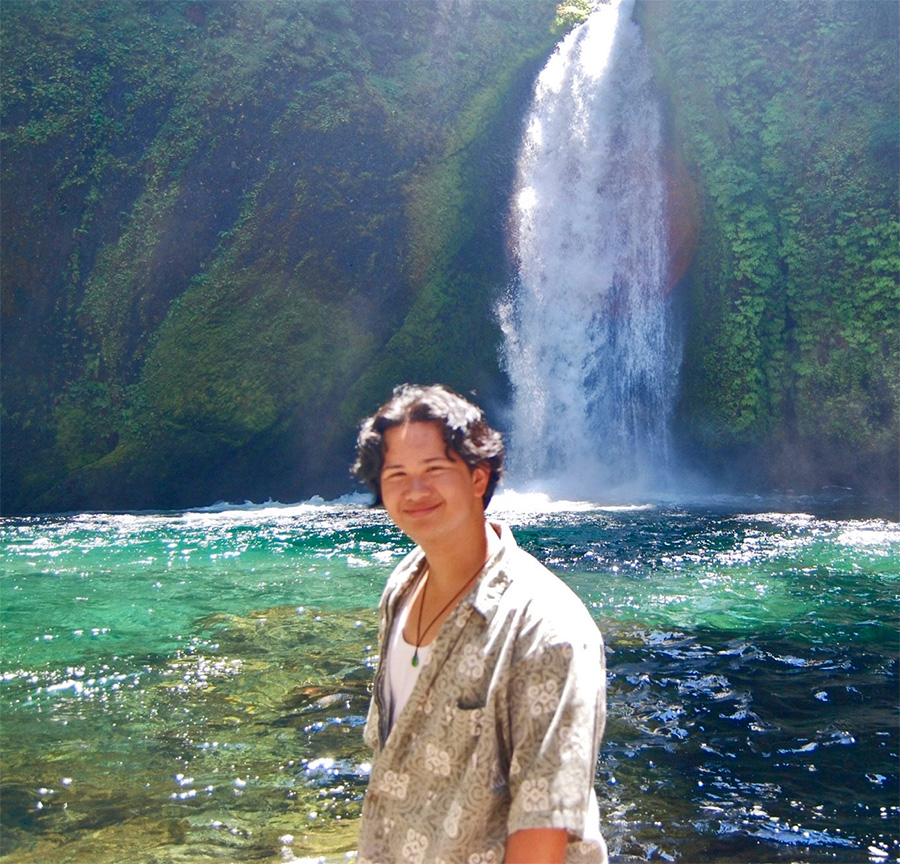 Photo of Jason Nuesa stanind in front a a large, jewel-green pool of water at the base of a tropical waterfall.