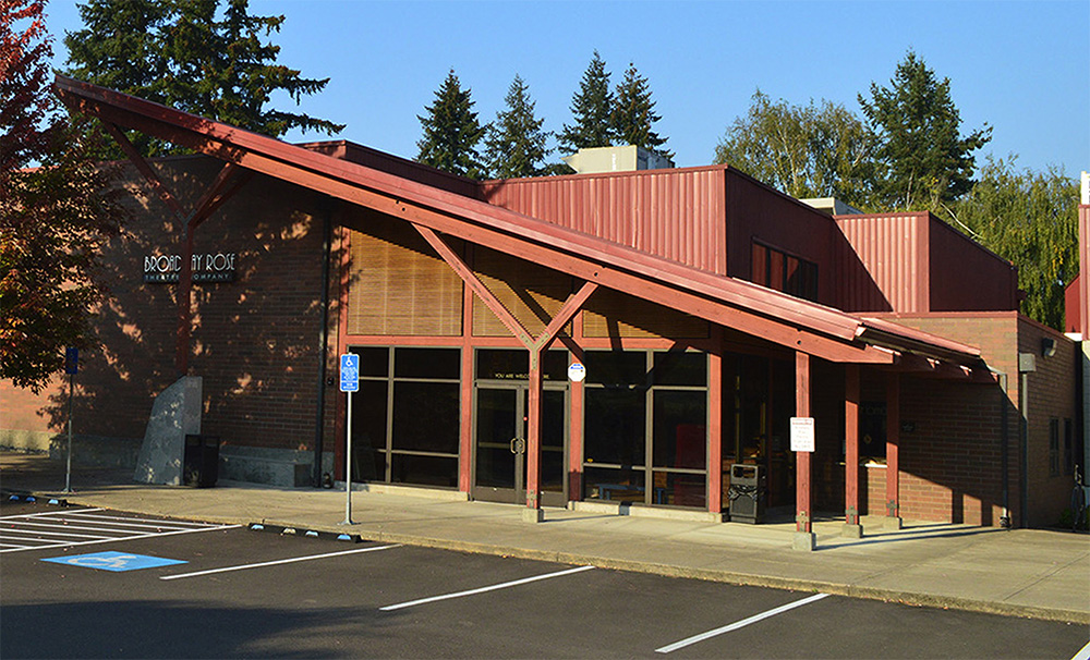 Photo of the exterior of the New Stage Theater. The entrance has a large, slanted awning over the front doors. The building is red brick with wood accents and a red roof.