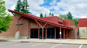 Photo of the front of the New Stage building. The roof over the atrium is sloped down and to the right. The front wall is all glass with a set of double doors leading into the building. The roof and support beams out front are red, but the building is mostly brown brick and beautiful wooden slats.