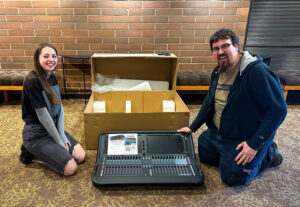 Photo of Brian Moen and Briana Fisher unboxing the new sound board last year, both are smiling big, very excited for this new piece of tech.